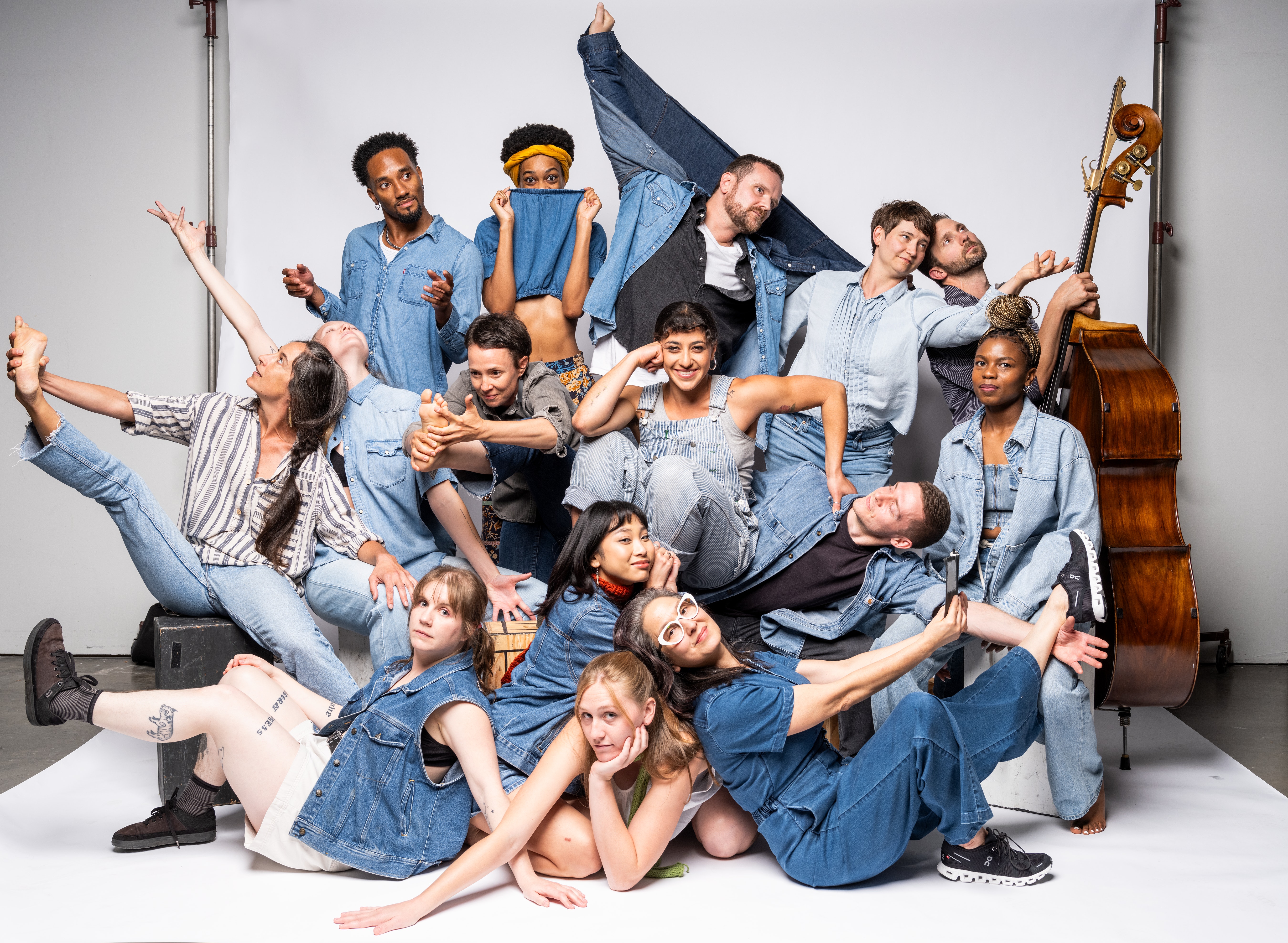 A group of several adults wearing various shades of blue denim are piled on a photo sweep, some smile at the viewer some are raising hands or legs.
