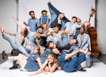 A group of several adults wearing various shades of blue denim are piled on a photo sweep, some smile at the viewer some are raising hands or legs.