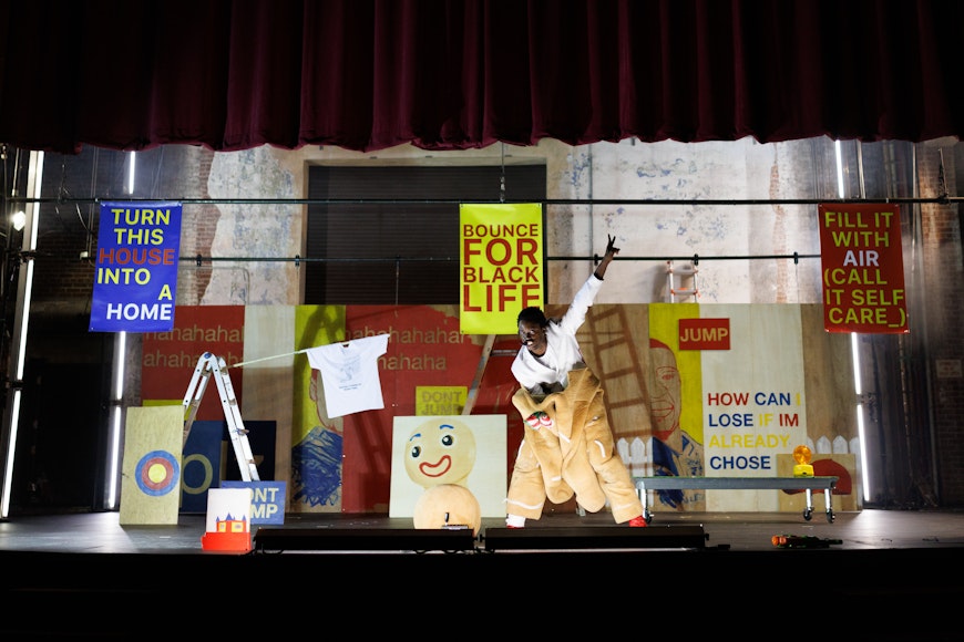 A dark-skinned man in a costume poses on stage with artwork in the background.