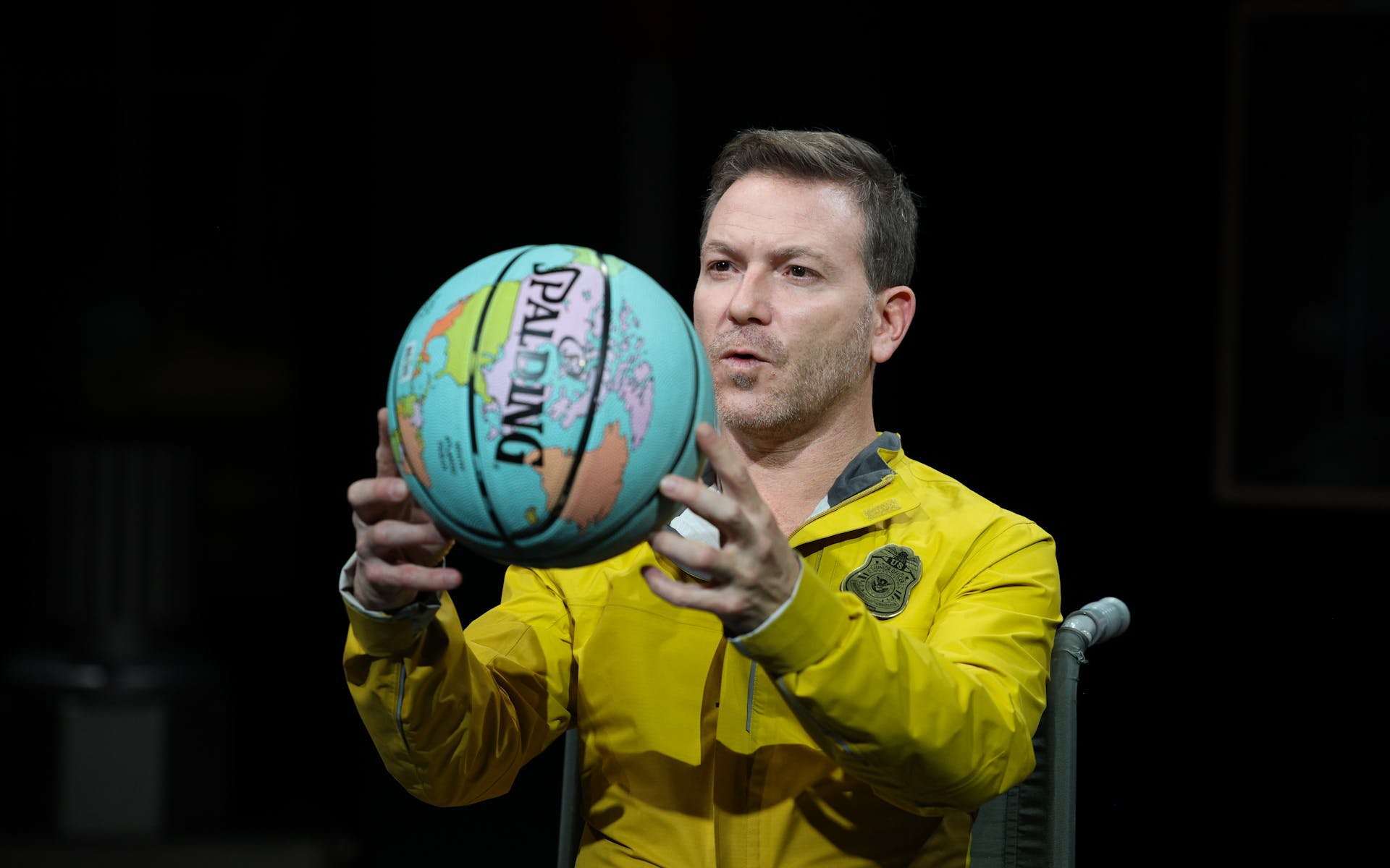 An actor in a bright yellow jacket holds up a basketball which features a print of the map of the world.