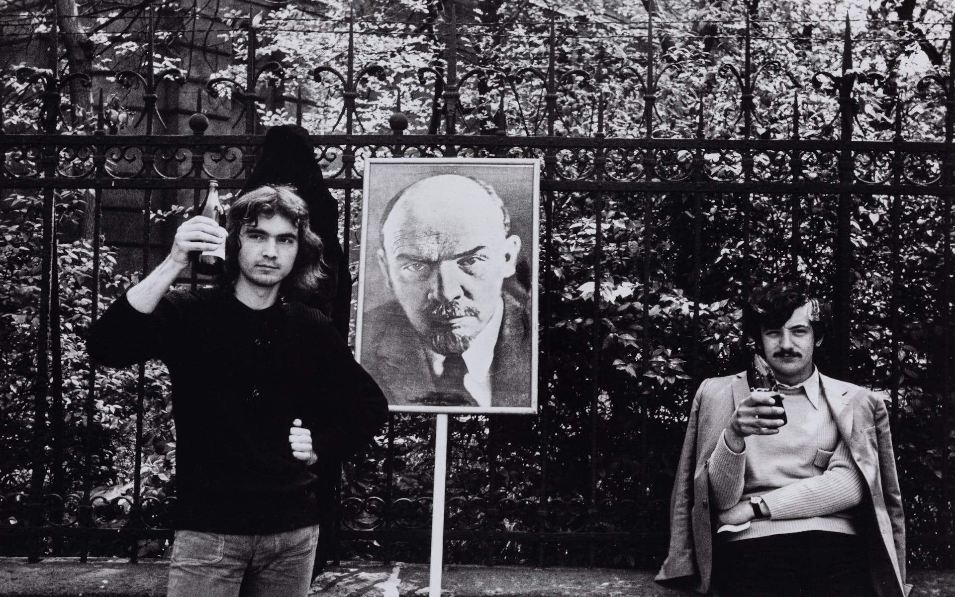 Black and white photograph of two men in the seventies leaning against a metal fence in front of a park with a large picture of Lenin attached to a wooden pole. Each man holds a bottle of beer and one of them lifts the bottle in his hand at the viewer.