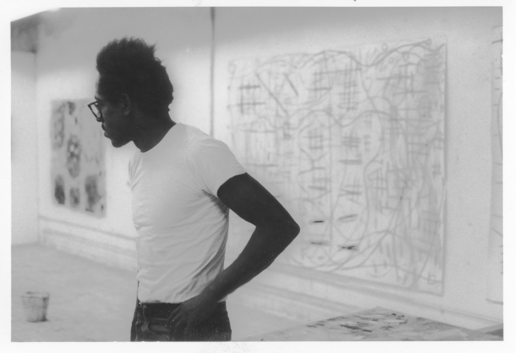 A young black man standing in front of large drawings hung on the wall.