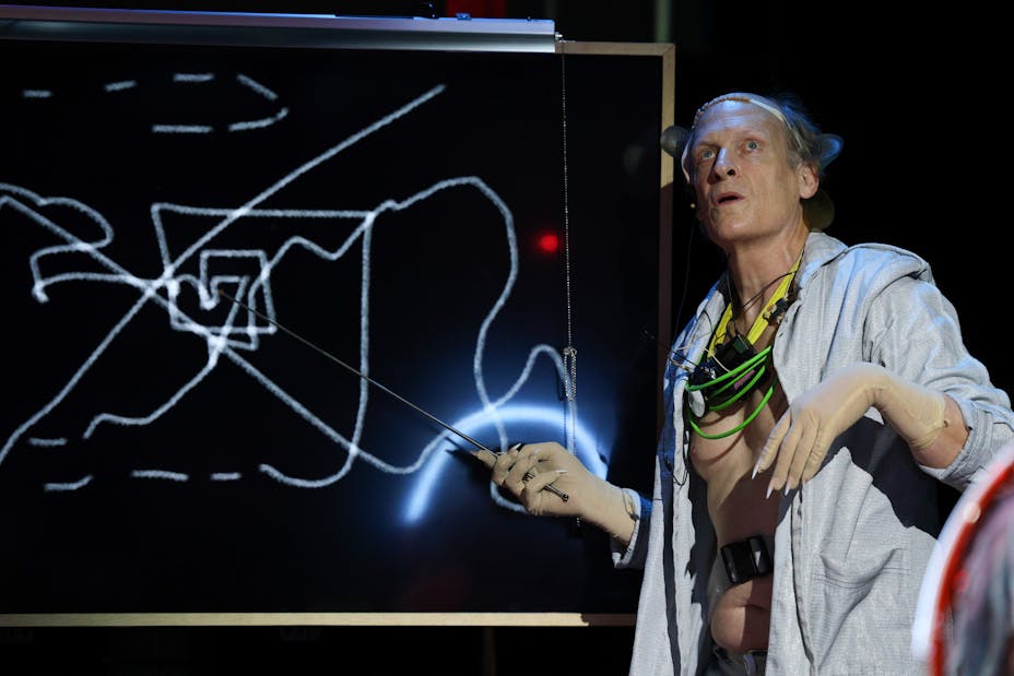 An older light-skinned man wearing wires points to a shape on a blackboard with a pointer.