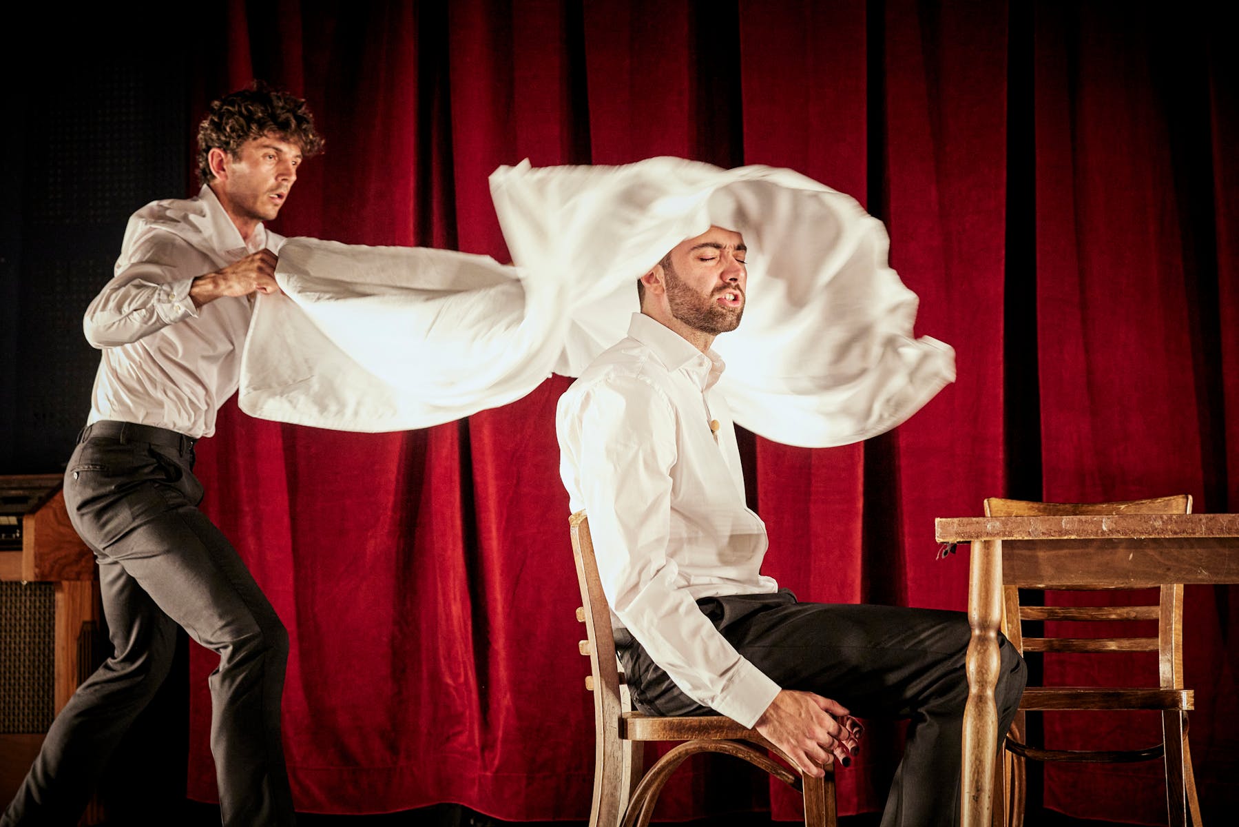 A light-skinned man throws a white sheet at a light-skinned man sitting down, against a red curtain.