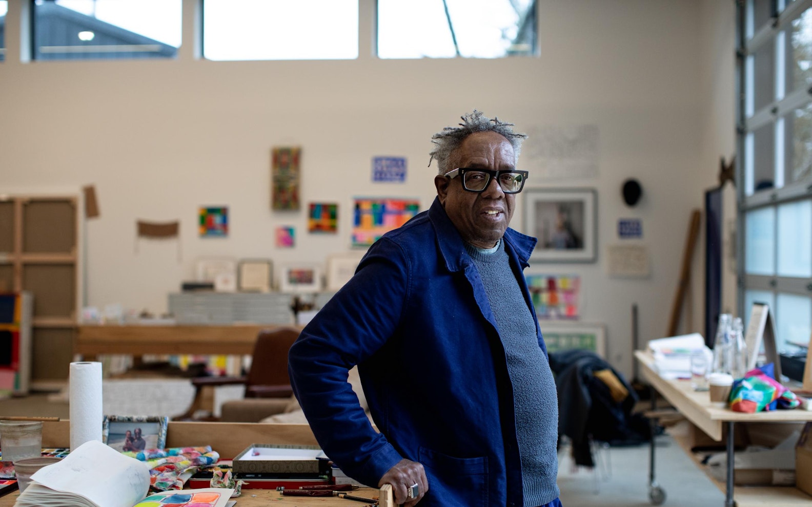 24. Portrait by Aundre Larrow An older black man wearing glasses and blue blazer, leaning against a table covered in crayons and sketchbooks.