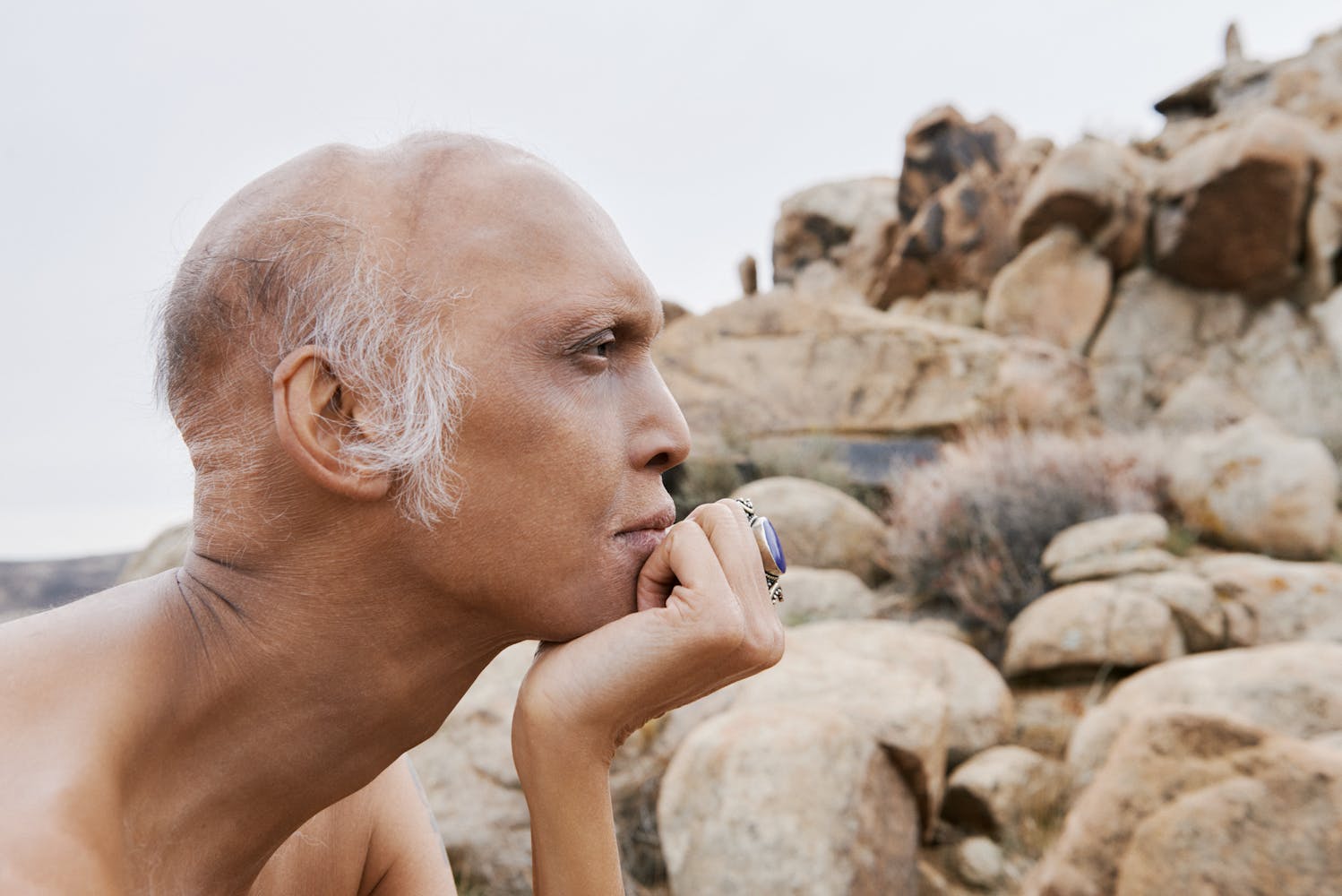 A person with medium skin rests their chin on their hand in front of a tan, rocky landscape.