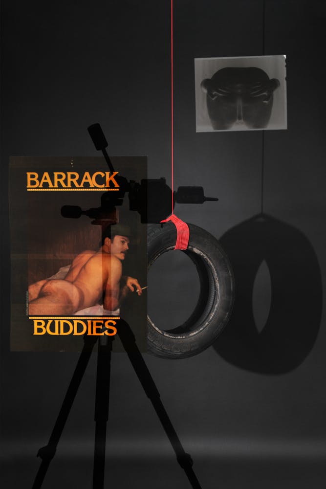 2. Photograph of an automobile tire suspended by a red rope against a black backdrop. A reflection of a camera on a tripod is prominent on the left, alongside a 4x5 film sheet of an anus and an image of a nude man on his stomach smoking a cigarette with the title “Barrack Buddies”.