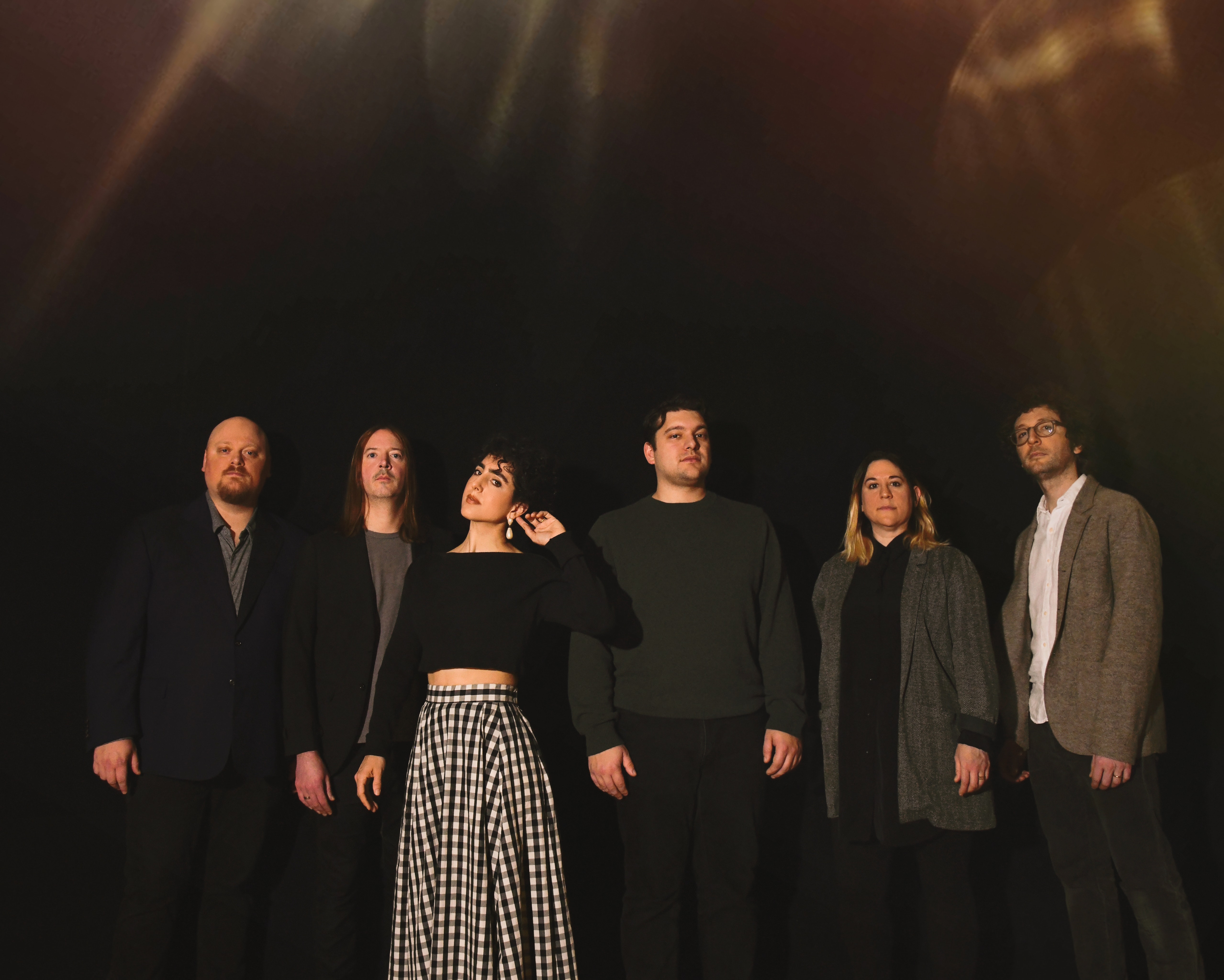 Six people in dark clothing stand and gaze at the viewer against a dark background.