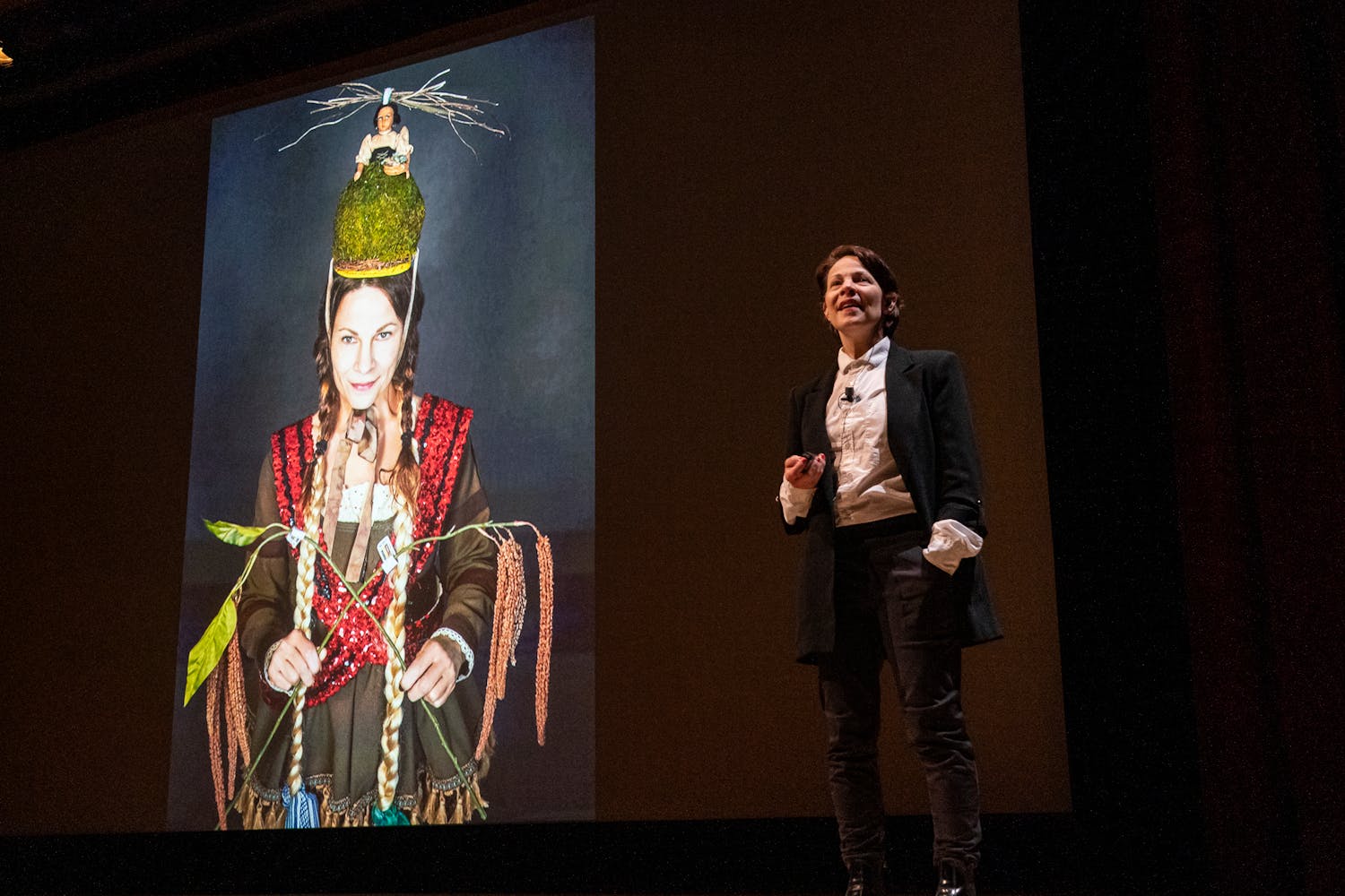 A person with light skin and short dark brown hair, wearing a suit coat and white button-up shirt, gives a lecture in front of an image of themselves wearing an ornate, vaguely medieval and somewhat silly costume.