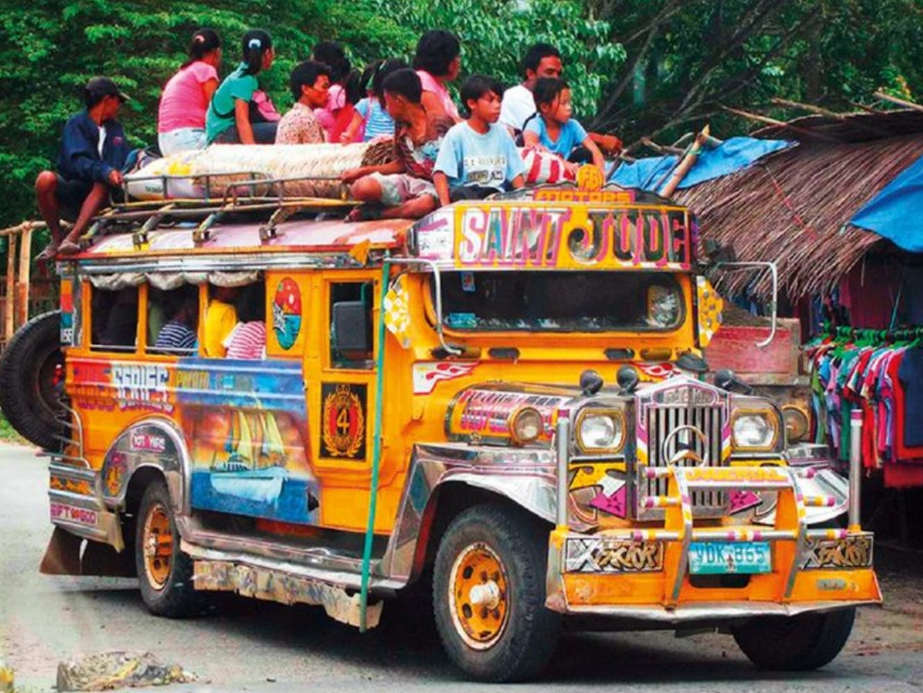 A bus painted with many images and colors drives down a jungle road with people riding inside and on top.