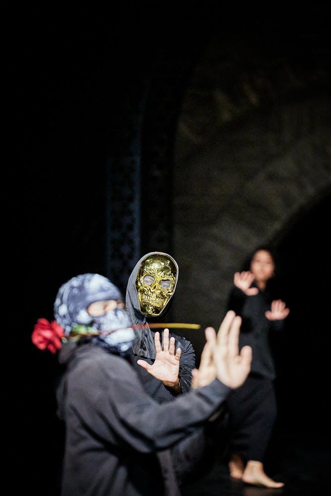 A dancer wearing a gold skull mask and a hooded sweatshirt watches another dancer in a hooded sweatshirt and skull mask hold a rose int heir mouth and lean back on stage.