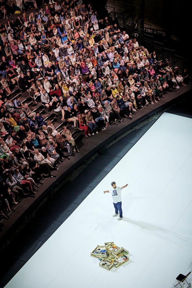 An overhead photograph of a performer with light skin on a white stage gesturing away from the audience.
