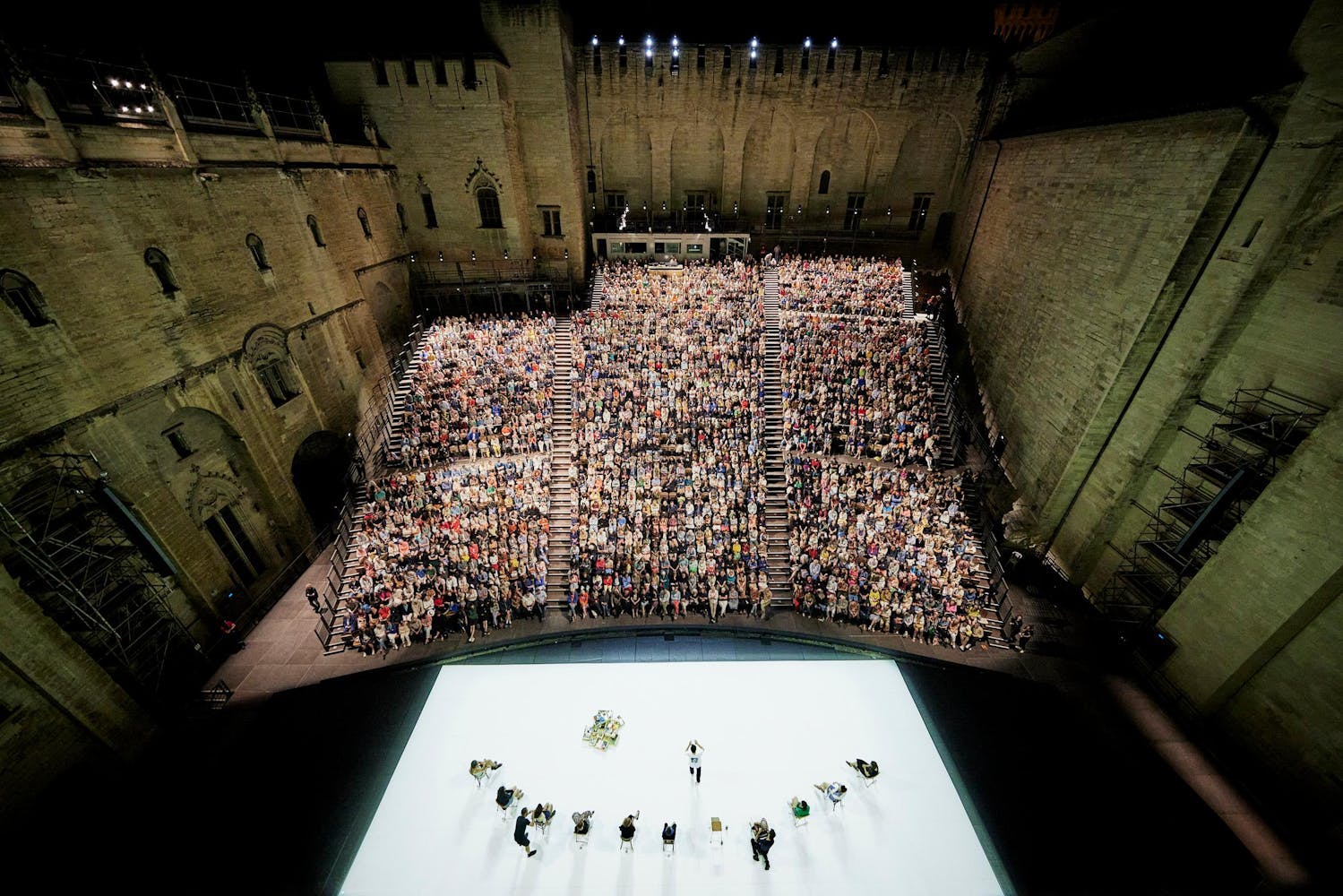 An overhead photograph of an ampitheater with a white rectangular stage and a large audience surrounded by old stone walls.