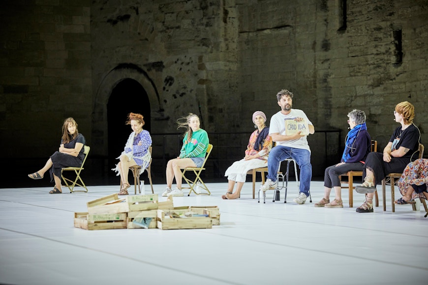 Seven people sit on-stage against a brick backdrop, with one person holding a book up that reads Candida.