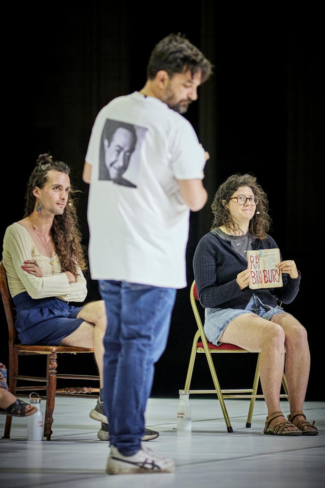 A seated performer with light skin holds open a book with 