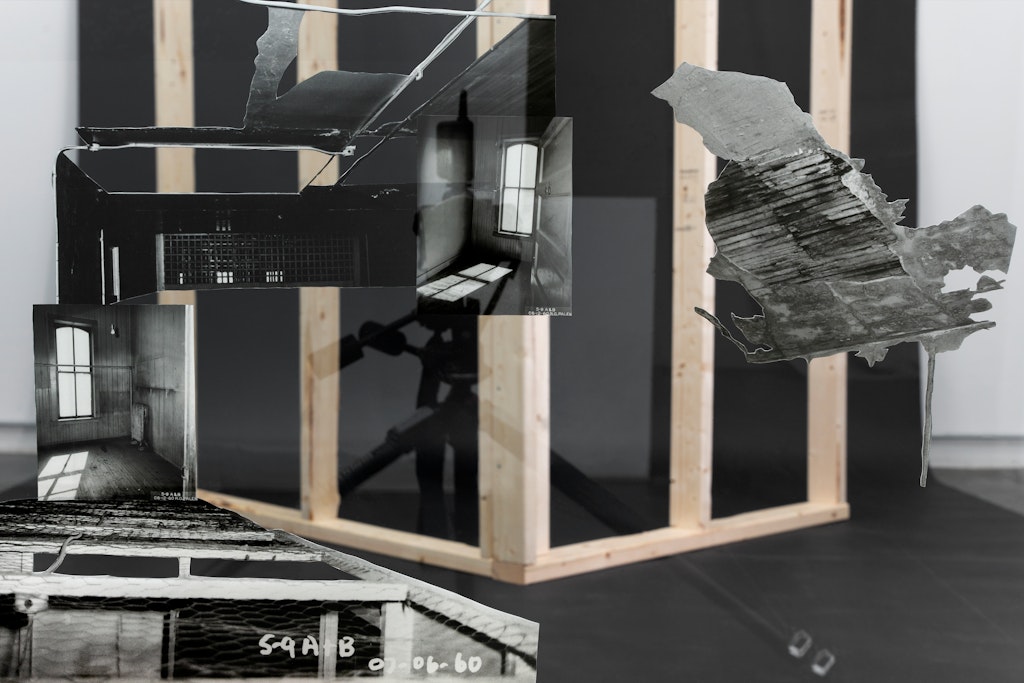 3. Photograph of a corner constructed using wooden studs against a black backdrop. A reflection of the camera is just off-center with multiple collage images from the Pioneer Hotel’s survey images before its demolition.
