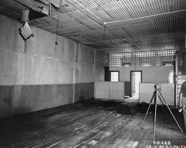 4. Archival image from a survey team by the City of Minneapolis of the Pioneer Hotel before its demolition. An empty tripod and a man carrying a camera take up the right side of the frame, among an otherwise empty and disheveled room.