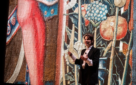 A person with light skin and short dark brown hair, wearing a suit coat and white button-up shirt, gives a lecture in front of a background of a detail of a medieval tapestry.