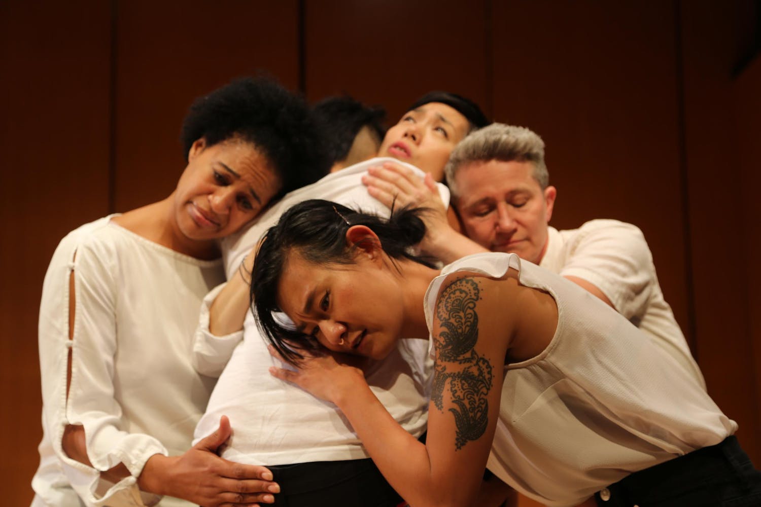 Five performers, all different skin tones and hair styles but all wearing white tops, embrace and console each other. The person in the front faces the camera and has a large paisley upper arm tattoo.