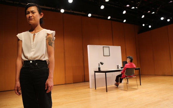 A performer with light/medium skin, dark hair pulled back, a white blouse and black jeans stands at the edge of the stage looking toward the audience. In the background, a performer sits on a chair facing away from the audience.