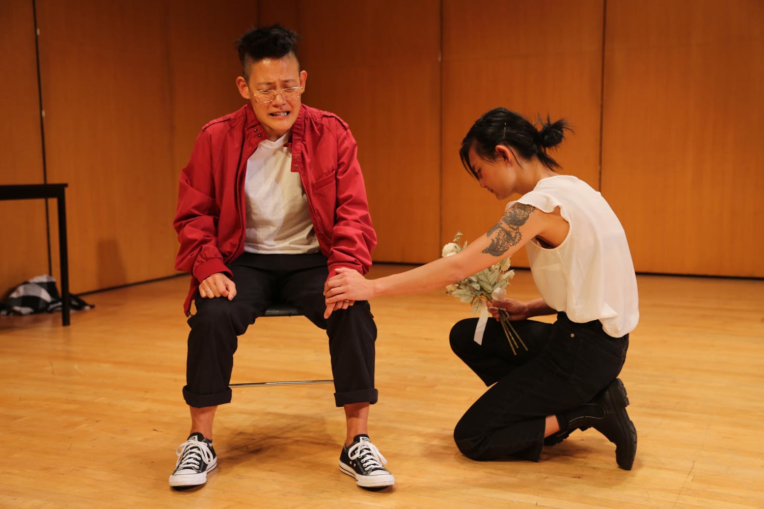 A performer with light/medium skin and short, spiky dark hair sits on a chair on a stage, wearing a red jacket and glasses. A second person with light skin, dark hair in a bun and a large upper arm tattoo kneels beside them, holding the other's hand as well as a bouquet of white flowers.