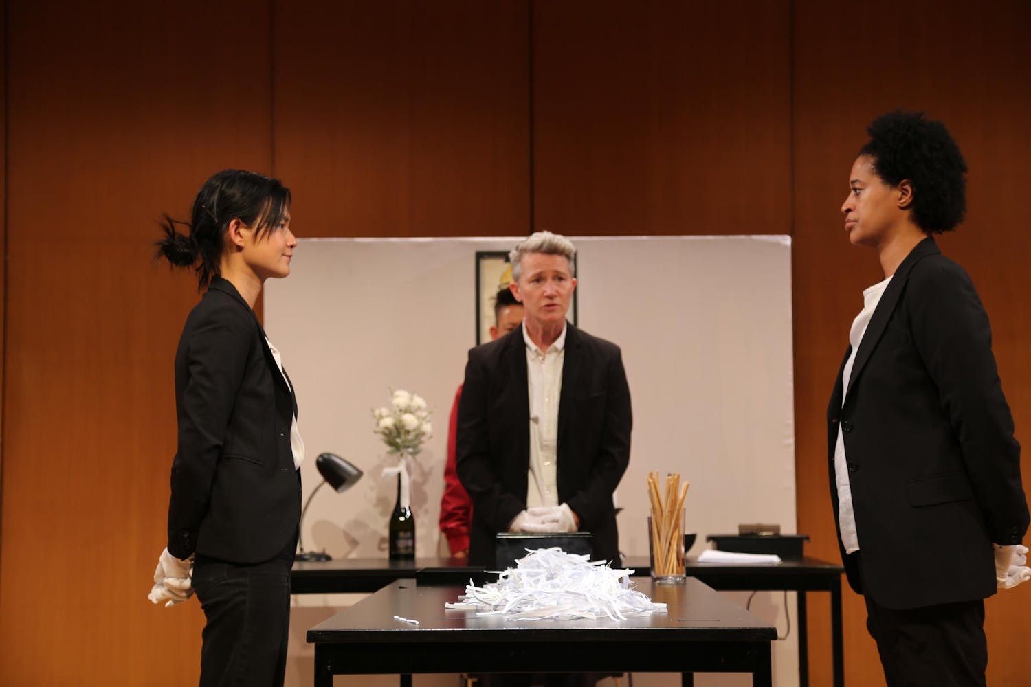 Two people stand facing each other, one with light/medium skin tone and dark hair in a bun, the other with medium/dark skin tone and a short afro. A person with light skin tone and short grey hair stands between them, behind a table. On the table lies a pile of white thin strips.