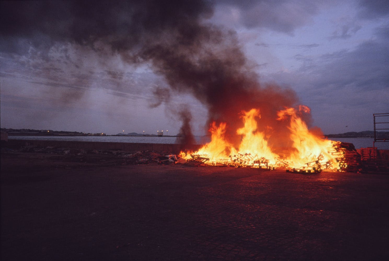 A pile of fire burns at dusk on land next to the ocean.
