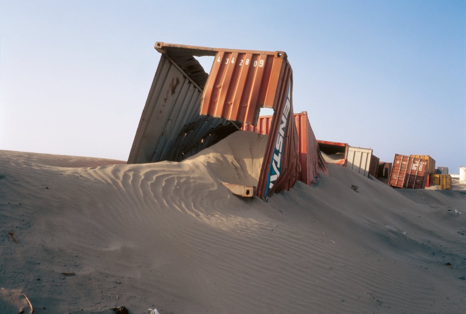 Abandoned shipping containers sit partially covered in sand dunes.