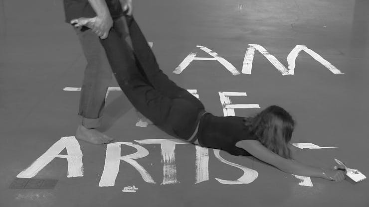 A person drags another person, who is holding a paintbrush, across the floor, which is painted with "I AM THE ARTIST" in block letters.