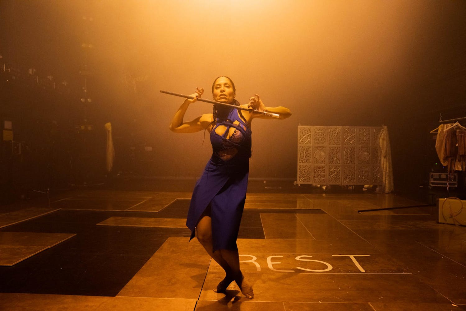 A person with dark skin, long black braids and a purple dress dances on a stage, holding a stick parallel to their shoulders. The word REST is written on the stage.