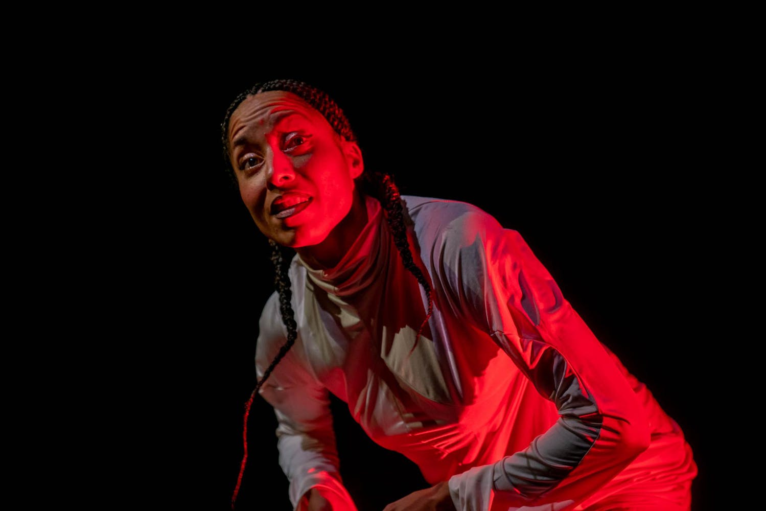 A person with dark skin, long dark braids, and a light turtleneck stands semi-hunched over on a red-lit stage.