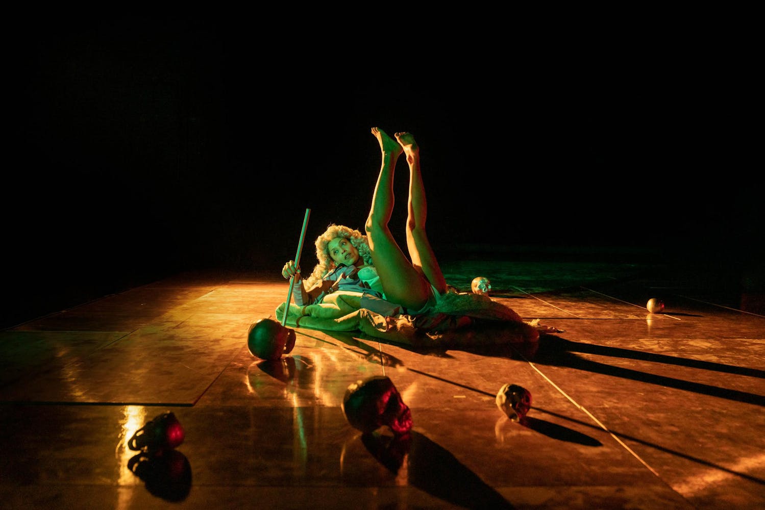 A performer with dark skin, wearing a colonial curly white wig, lays on a stage with both legs lifted, holding a bar. Several skulls are scattered on the stage.
