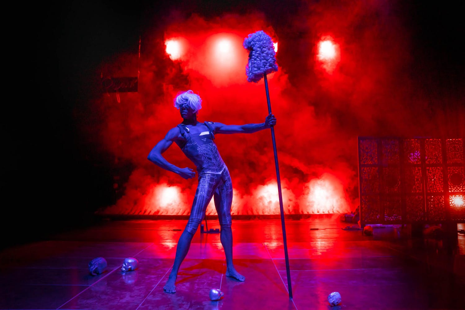 A performer wearing a short white wig stands flexing their muscles triumphantly on a red, foggy backlit stage. They hold a tall stick with a colonial wig on the top. Several skulls are on the ground.