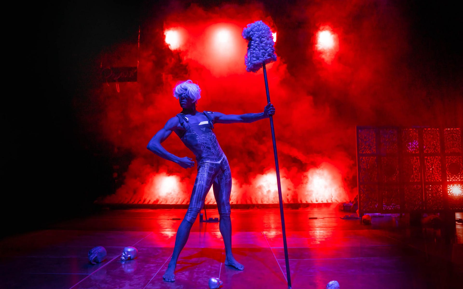 A performer wearing a short white wig stands flexing their muscles triumphantly on a red, foggy backlit stage. They hold a tall stick with a colonial wig on the top. Several skulls are on the ground.