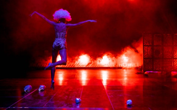 A performer dances on a dramatically lit, foggy stage of reds and purple. The dancer is facing away from the audience and wearing a white puffy wig. Four silver skulls lay on the stage.