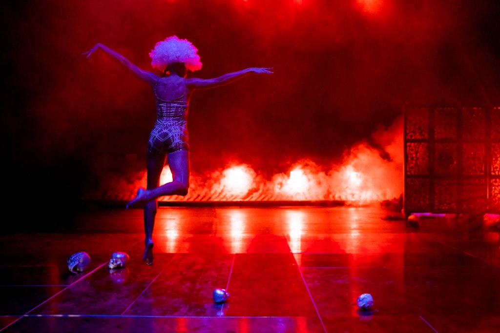 A performer dances on a dramatically lit, foggy stage of reds and purple. The dancer is facing away from the audience and wearing a white puffy wig. Four silver skulls lay on the stage.