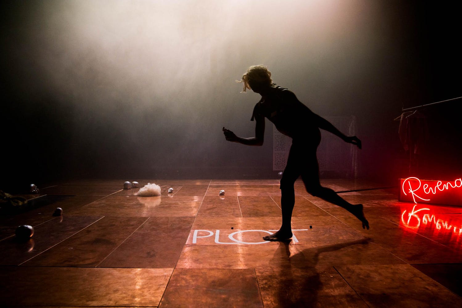 A silhouette of a moving performer standing on the word PLOT written on the stage; part of a word in neon is lit on the right 