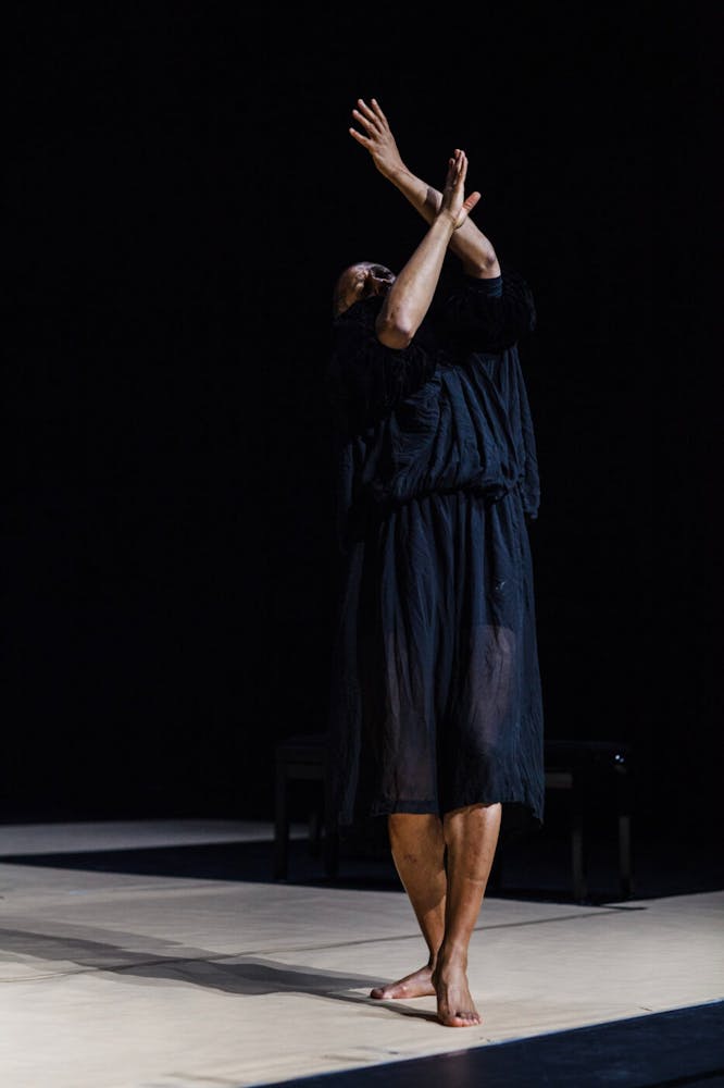 A dance with dark skin wears a black tunic and holds her arms up over their head.
