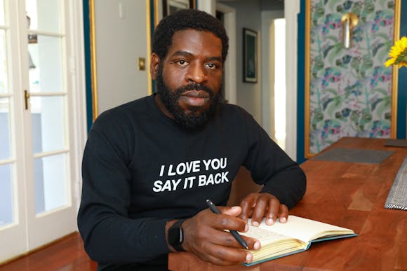 A man with dark skin holds a notebook and a pen while sitting at a table indoors and looking at the viewer. He wears a sweartshirt with the words "I Love You Say It Back."