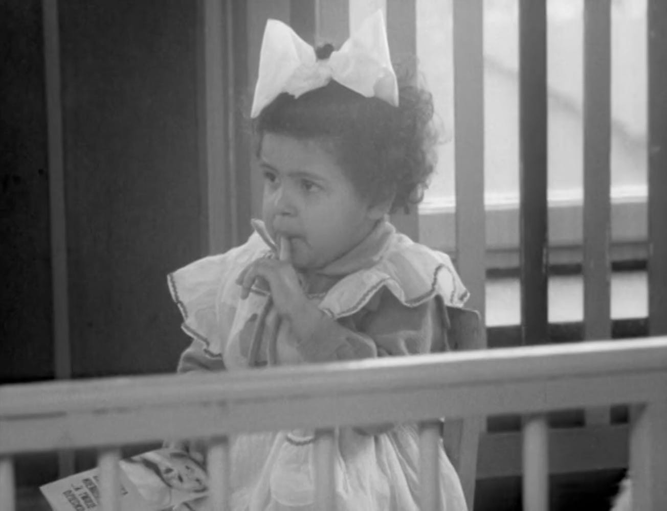 A young girl with dark skin and hair chews on her finger while wearing a dress and bow in her hair.
