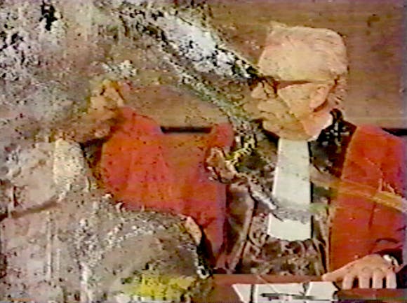 A man in a red jacket sits at a desk. The film emulsion on the left side of the image is damaged.