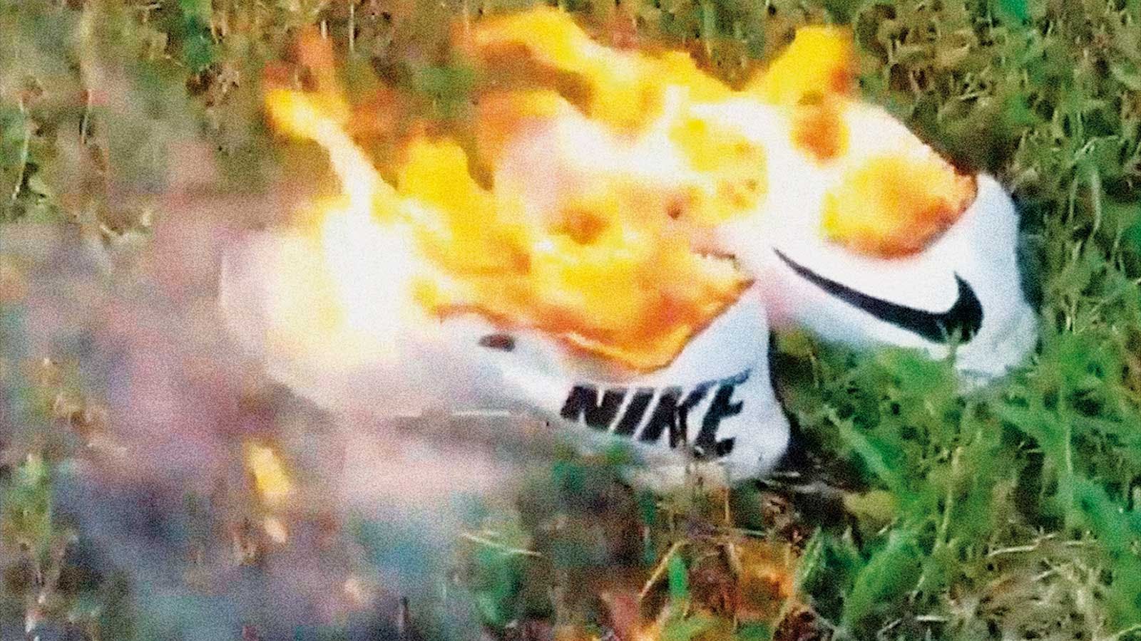 A pair of white Nikes on fire in the grass, blurred slightly from movement and smoke.