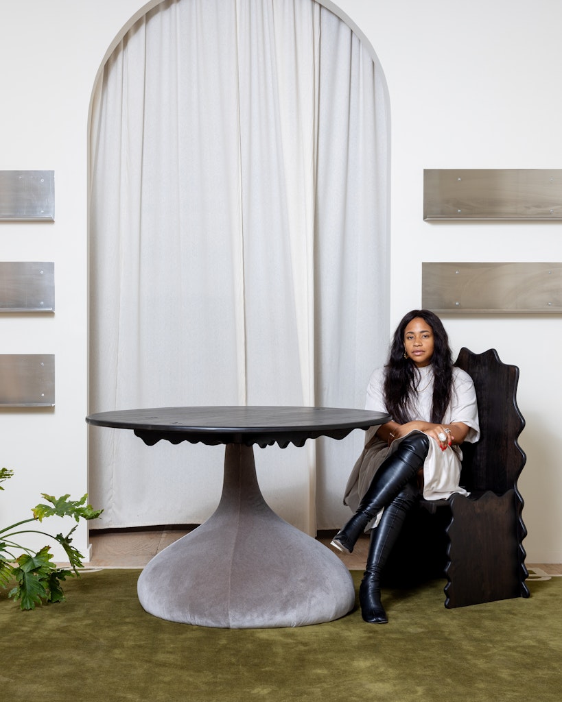A woman with dark skin and long dark hair sits on a wavy wooden chair next to a wavey wooden table and looks at the viewer.
