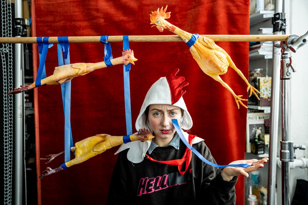 A performer with light skin poses with strips of blue painters tape and yellow rubber chickens in front of a red backdrop.