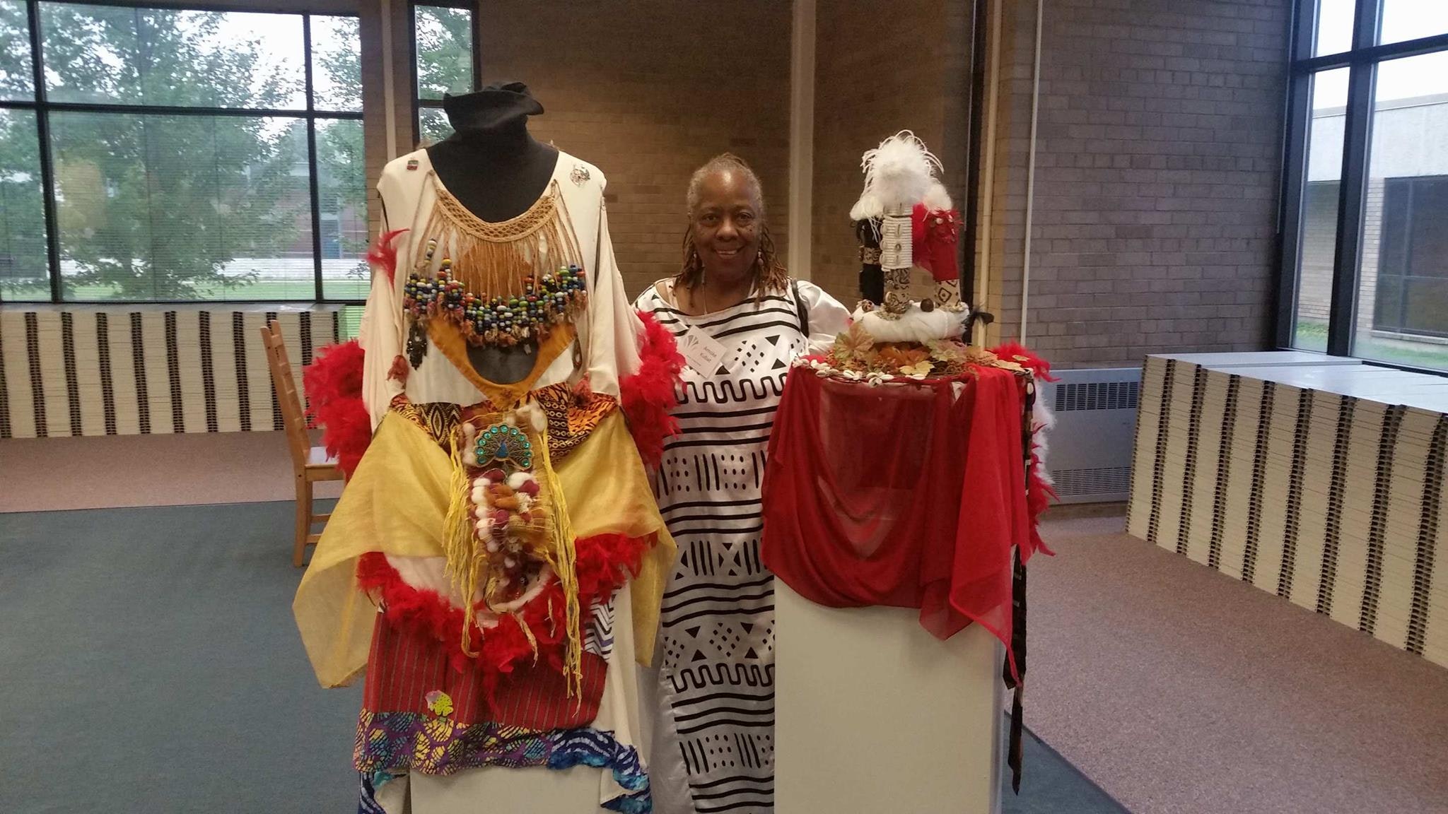 A dark skinned woman standing and smiling between a costumed dress form and a sculpture on pedestal. The dress form's clothes and sculpture have many layers of fabric and different colors and textures. 