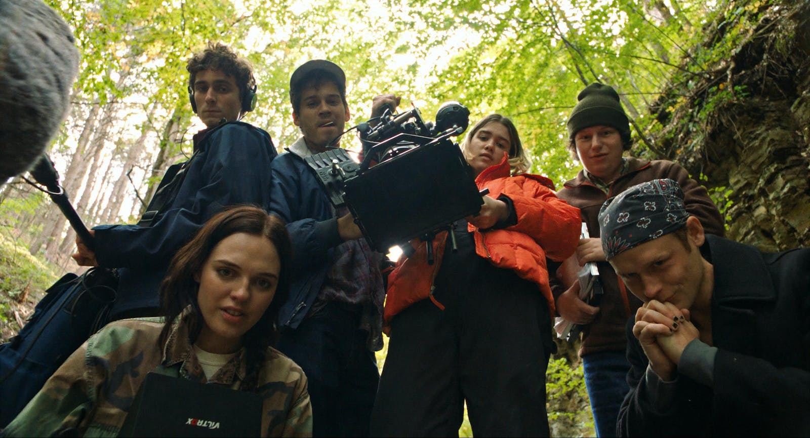 A film crew of six young people with equipment standing and crouching, looking directly at us. They are outdoors in woods.