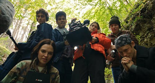 A film crew of six young people with equipment standing and crouching, looking directly at us. They are outdoors in woods.