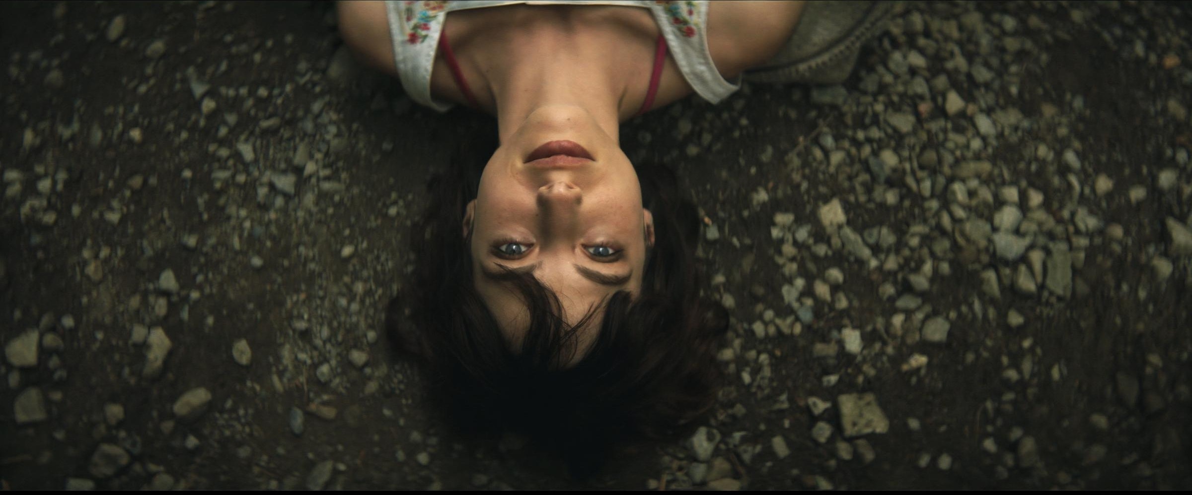 Upside down head-and-shoulders shot looking down at a woman with brown hair and bangs, laying on a dirt and gravel.