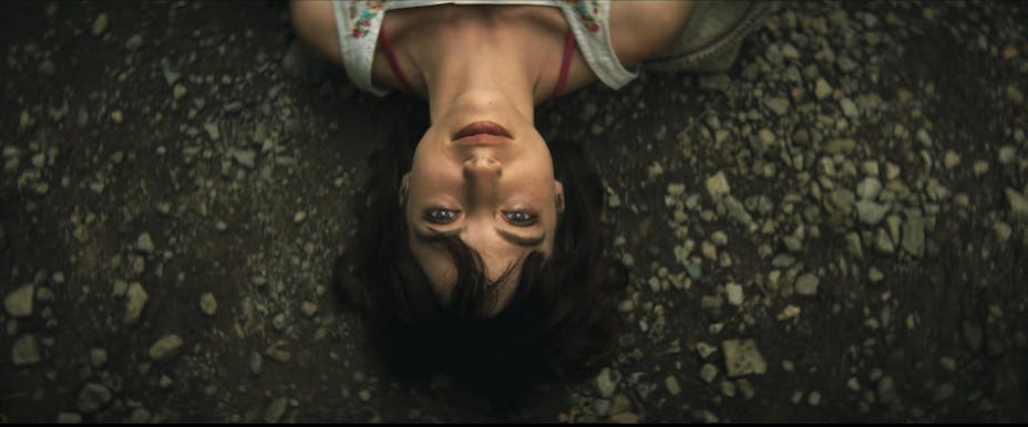 Upside down head-and-shoulders shot looking down at a woman with brown hair and bangs, laying on a dirt and gravel.