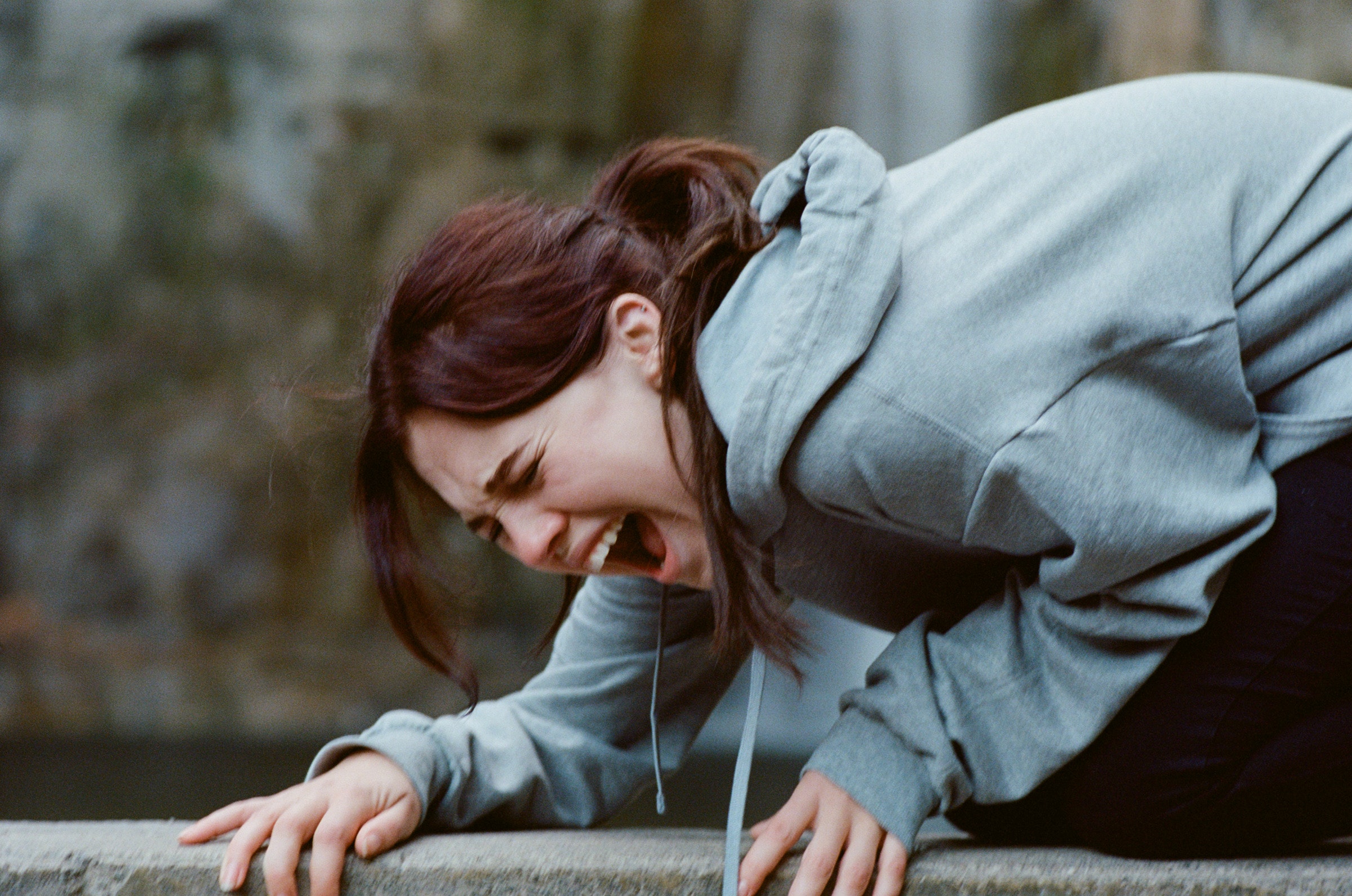 Still of a woman with light skin, brown hair in a ponytail, and a gray hoodie kneeling, crouched over and screaming.